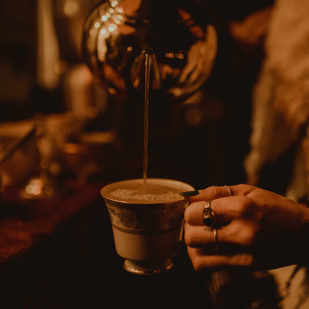 A hand adorned with rings delicately holds an ornate teacup while a hot beverage is being poured from a brass teapot, set against the warm, inviting backdrop of an exclusive Club Magic Hour private tea ceremony. The scene exudes a cozy, vintage atmosphere and resonates with the elegance embodied in the Founding Angel Guide: $5,000 Pledge.