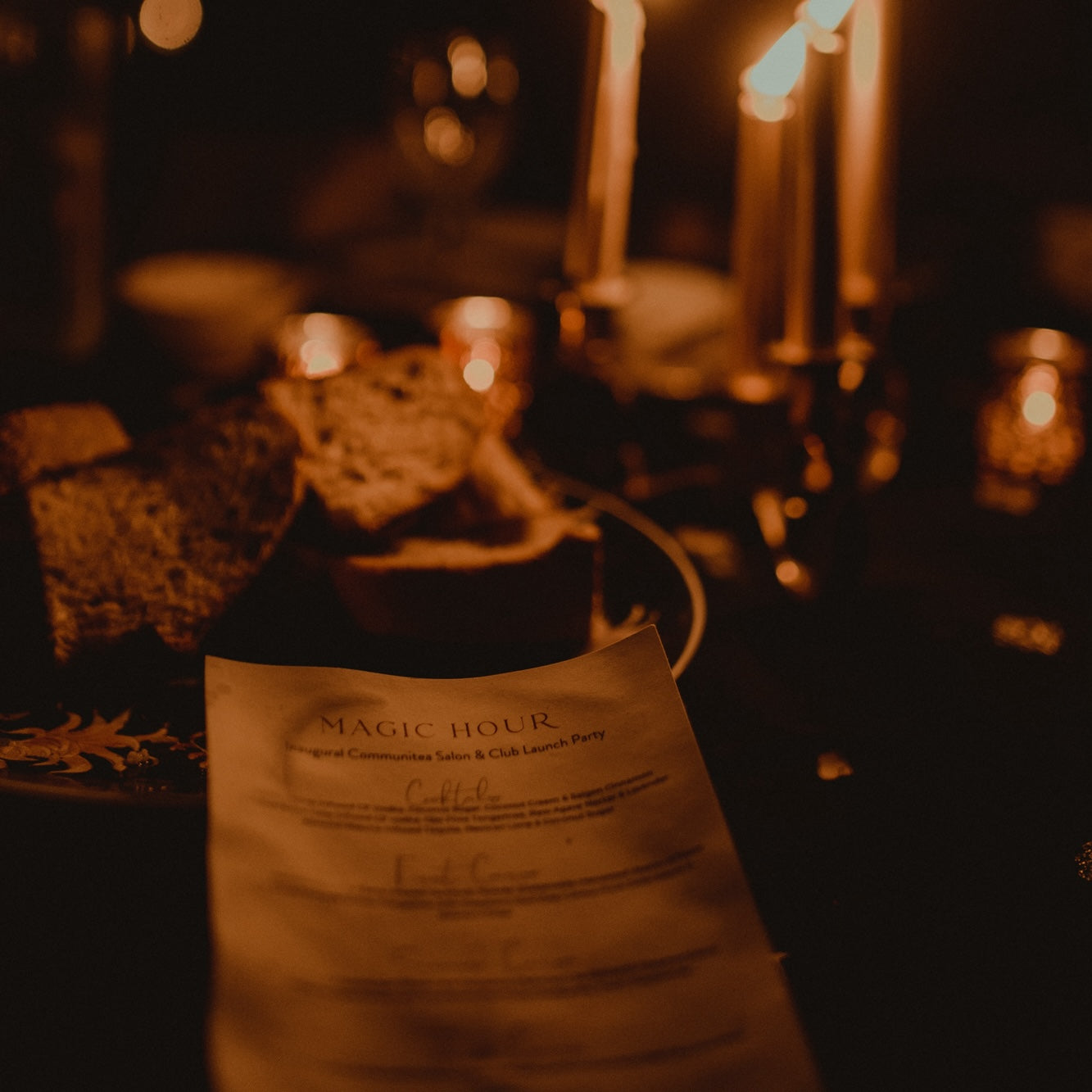 A dimly lit table set with candles and a plate of bread. In focus is a menu titled "Archangel of Magic Hour" featuring details about an exclusive private event for those who pledge $10,000. The overall ambiance is warm and intimate, with flickering candlelight adding a cozy atmosphere for legacy supporters.