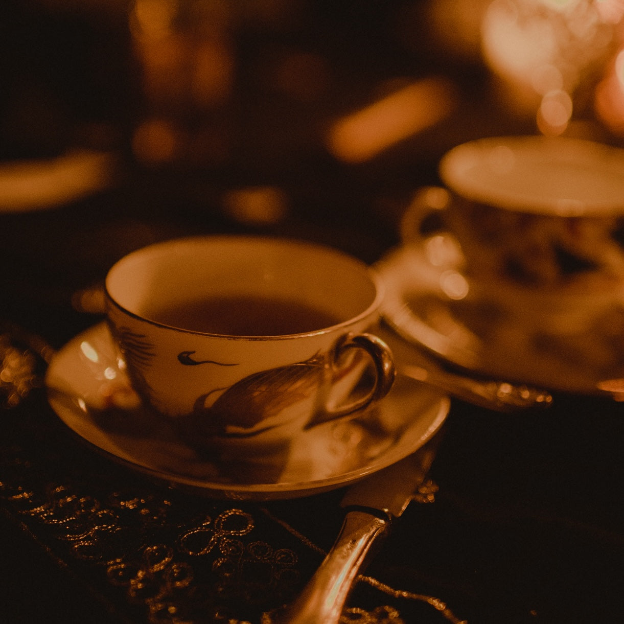 A close-up of an ornate teacup filled with tea, resting on a saucer. Another similar teacup is partially visible in the background. The warm, dim lighting gives a cozy, inviting ambiance to the scene, evoking the exclusive and luxurious experience promised by Club Magic Hour's Miracle Maker: $1,000 Pledge.