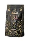 A black tea bag called "The World" by Magic Hour displays a colorful abstract figure and floral motifs in its intricate design. The packaging is decorated with stars and white flowers.