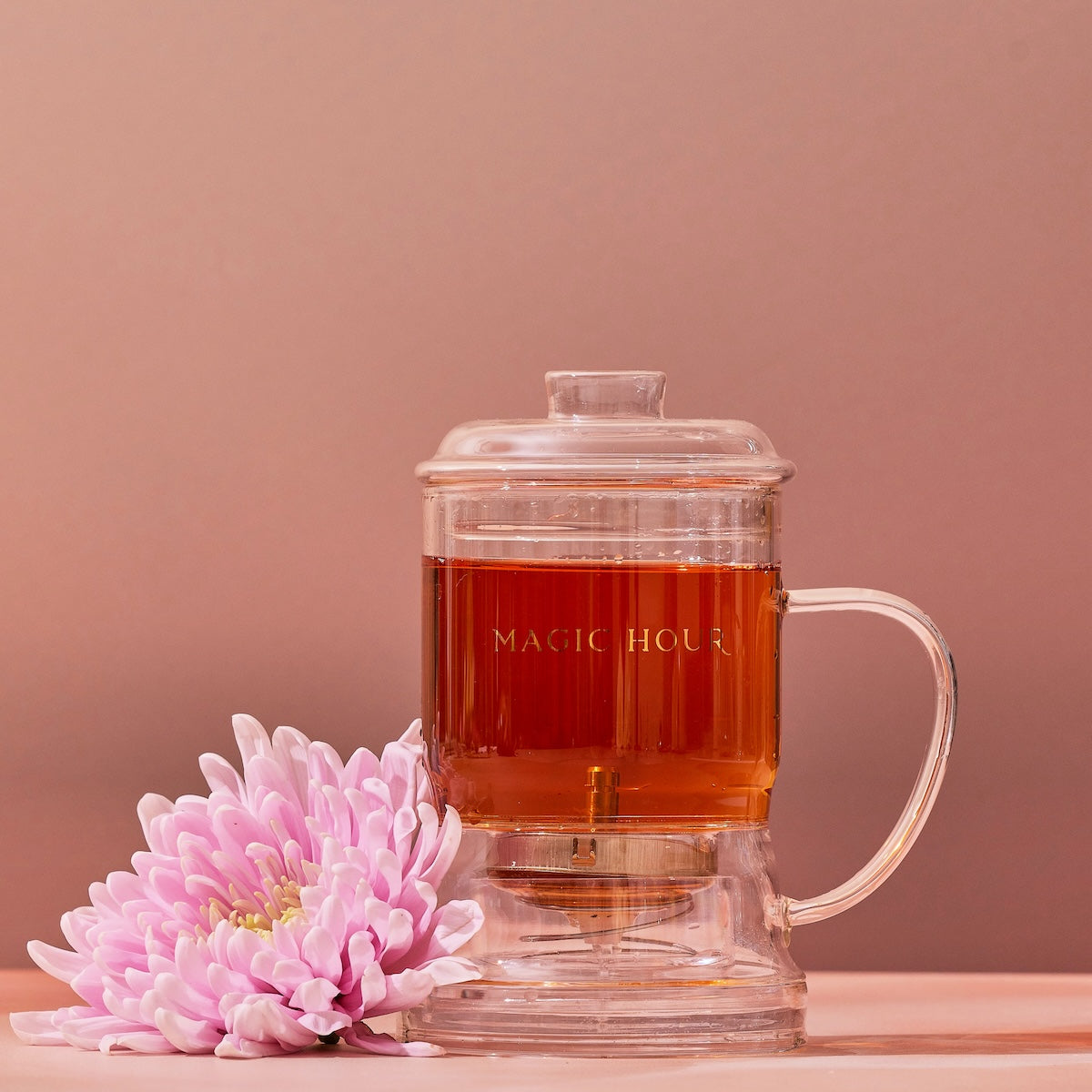 A  Glass Tea Infuser, elegantly designed and currently filled with tea, rests on a light wooden surface against a muted pink background. A pink chrysanthemum flower lies beside the infuser, adding a delicate touch to the composition.