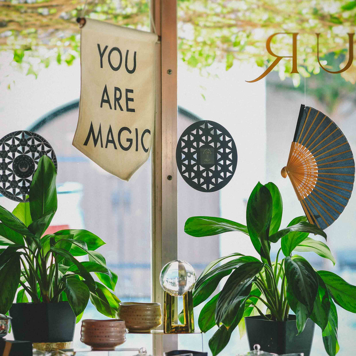 A You are Magic Flag hangs in a window beside two potted plants, a decorative fan, and geometric patterns—an inspiring wall decor piece with sunlight filtering through leaves outside.