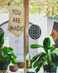 A You are Magic Flag hangs in a window beside two potted plants, a decorative fan, and geometric patterns—an inspiring wall decor piece with sunlight filtering through leaves outside.