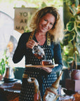 A woman pours tea from a glass teapot into a cup at Ojai Tea Salon, surrounded by plants and decor, wearing a dark apron with gold accents. Scene showcases Tea Essentials + In Person & Virtual Tea School Course.