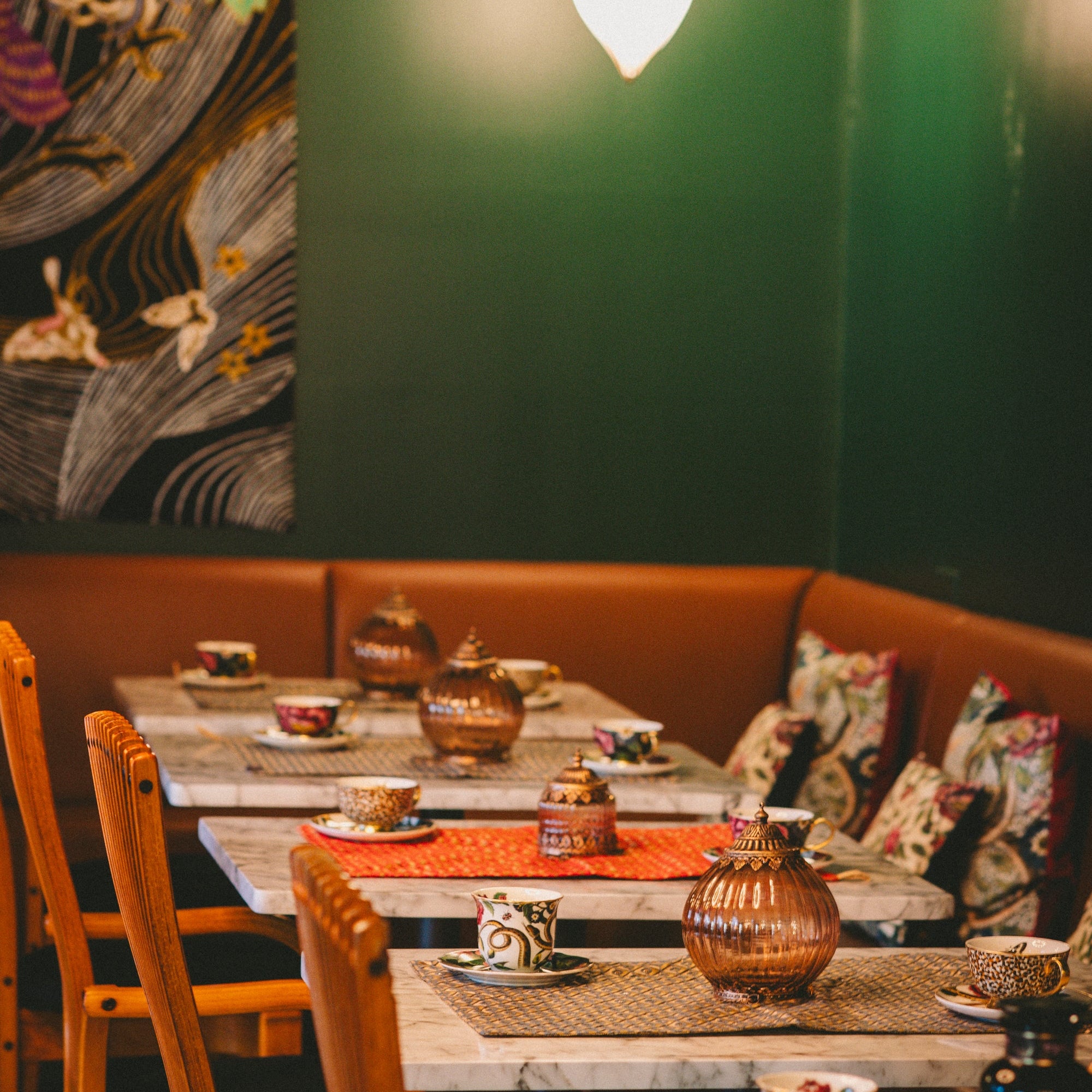 A cozy Ojai Tea Salon with wooden chairs, marble tables featuring Tea Essentials + In Person &amp; Virtual Tea School Course, patterned cups, colorful cushions on a curved bench, warm lights, and a floral mural on the green wall.