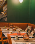 A cozy Ojai Tea Salon with wooden chairs, marble tables featuring Tea Essentials + In Person & Virtual Tea School Course, patterned cups, colorful cushions on a curved bench, warm lights, and a floral mural on the green wall.