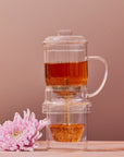 An elegant design, the clear Glass Tea Infuser by Alibaba includes a lid and sits atop the matching glass Magic Hour Glass Tea Infuser, allowing tea to seep from the pot above into a lower container. To the left of this premium quality setup, a pink chrysanthemum flower rests on the surface. The background is a warm, light brown color.