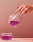 A hand wearing rings pours vibrant purple tea from a clear Magic Hour Glass Teapot into a matching glass teacup against a soft, peach-toned background.