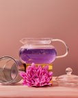 A handcrafted 450ml Magic Hour Glass Teapot filled with purple liquid is set on a wooden surface. Beside it, there is a metal mesh tea infuser, a vibrant pink flower, and a glass lid. The background gradient transitions from peach to mauve, complementing the elegant setup.