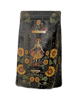 A vibrant black package adorned with detailed illustrations of sunflowers and leafy vines, featuring a central image of a woman in a flowing dress amidst sunflowers. Text reads "Magic Hour" and "The Sun: Energizing Adaptogenic Herbal Tea with Yaupon, Moringa & Turmeric," hinting at the natural blend within.