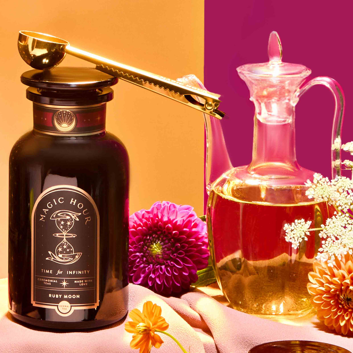 A dark jar labeled “Magic Hour” is paired with the Perfect Cup Tea Scoop with Pouch Clip in gleaming gold, resting beside a glass decanter of liquid and surrounded by colorful flowers on a pink and orange background—an elegant scene showcasing exquisite tea accessories.