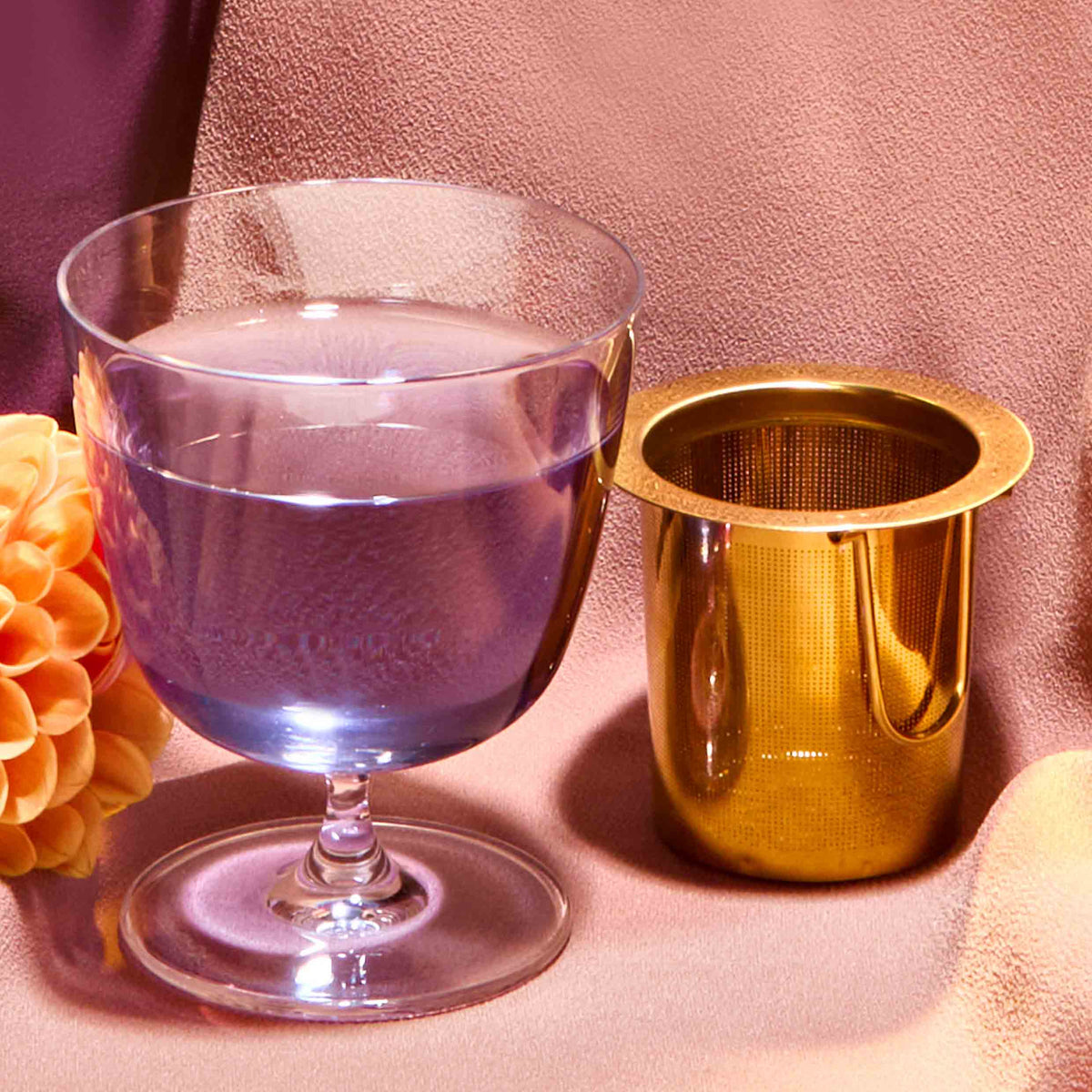 A clear glass of water stands next to the Midas Touch: Golden-Hued Tea Strainer cup on a textured, light pink surface with a peach-colored flower nearby.
