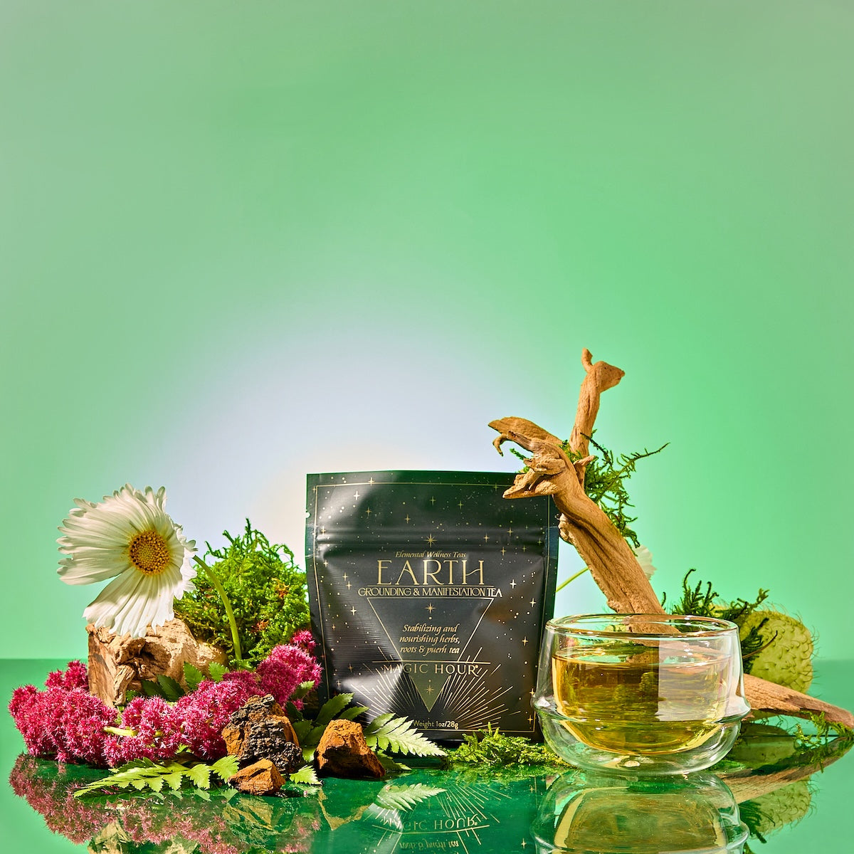 A packet labeled "Earth: Stabilizing and Nourishing Herbs, Roots, and Puerh tea" rests among moss, flowers, and a wooden stick, with a glass cup of grounding tea in front. The green gradient background enhances the calming atmosphere.