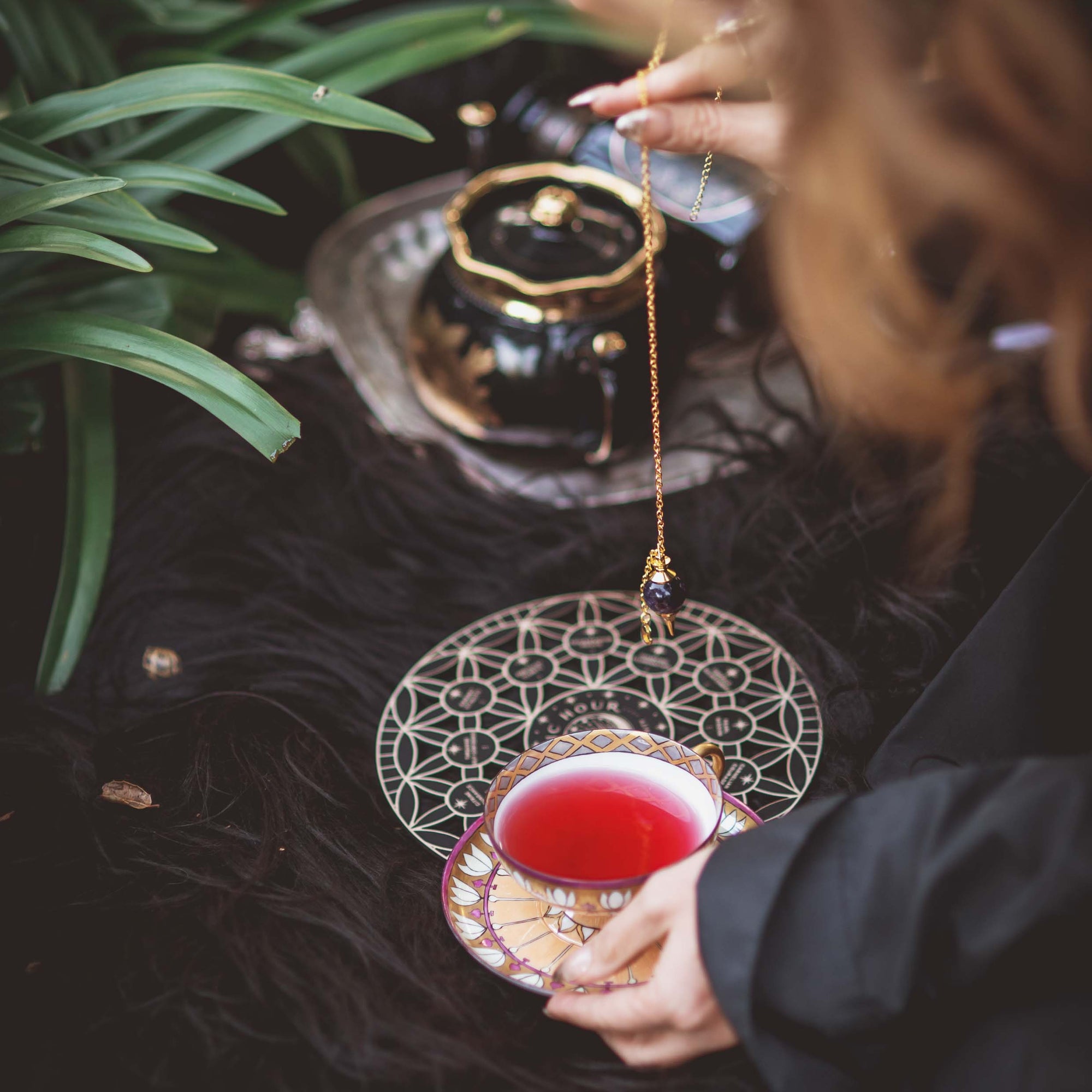 A person holds a cup of red tea beside the Crystal Grid: Flower of Life Tea Ceremony Altar, with greenery in the background. A crystal pendulum is suspended above the grid, evoking a ritual or meditation focused on intentional manifestation.