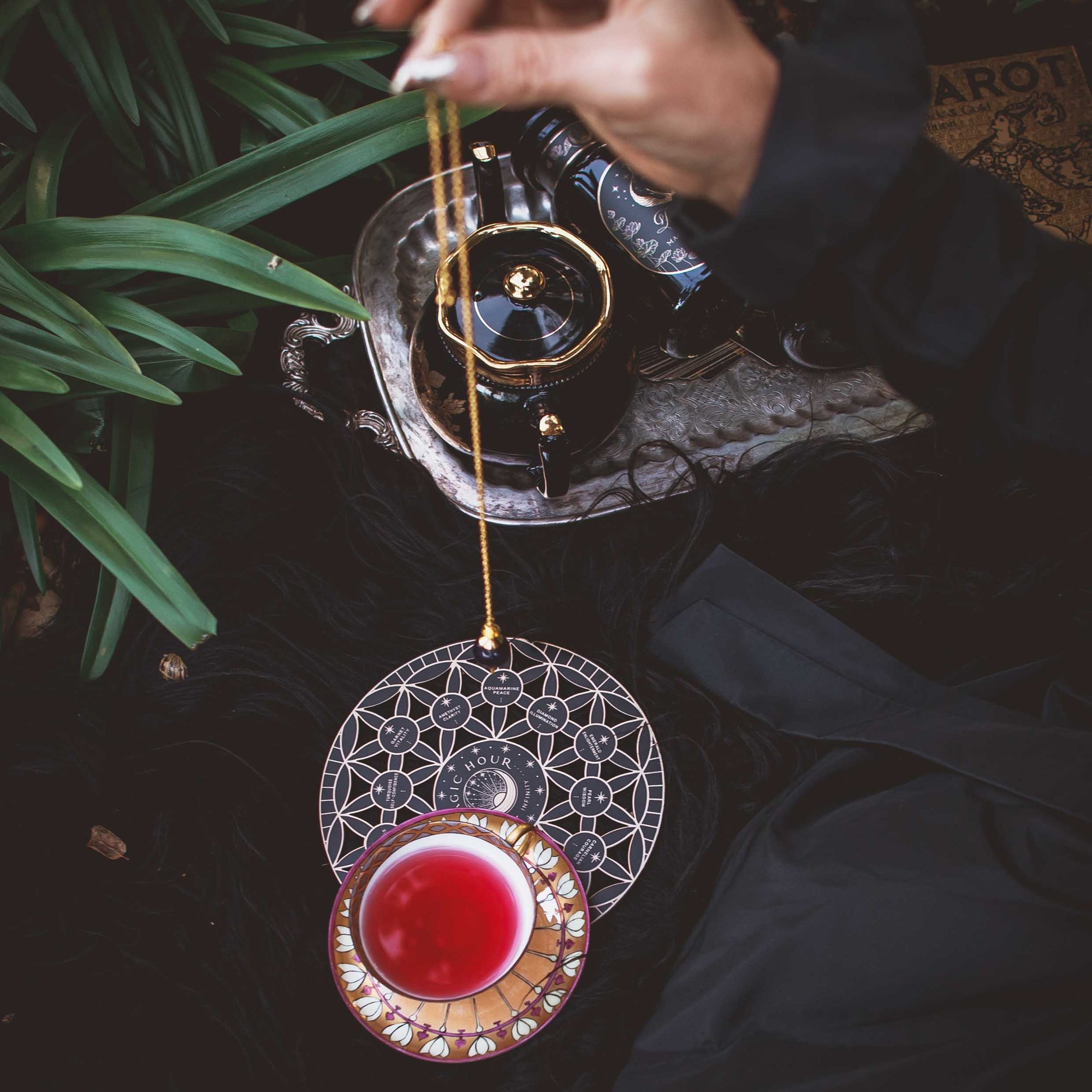 A hand holds a pendulum above the Crystal Grid: Flower of Life Tea Ceremony Altar, surrounded by red tea in an ornate cup, a teapot, and green leaves, evoking a mystical, tarot-inspired setting for ritual tea ceremonies.