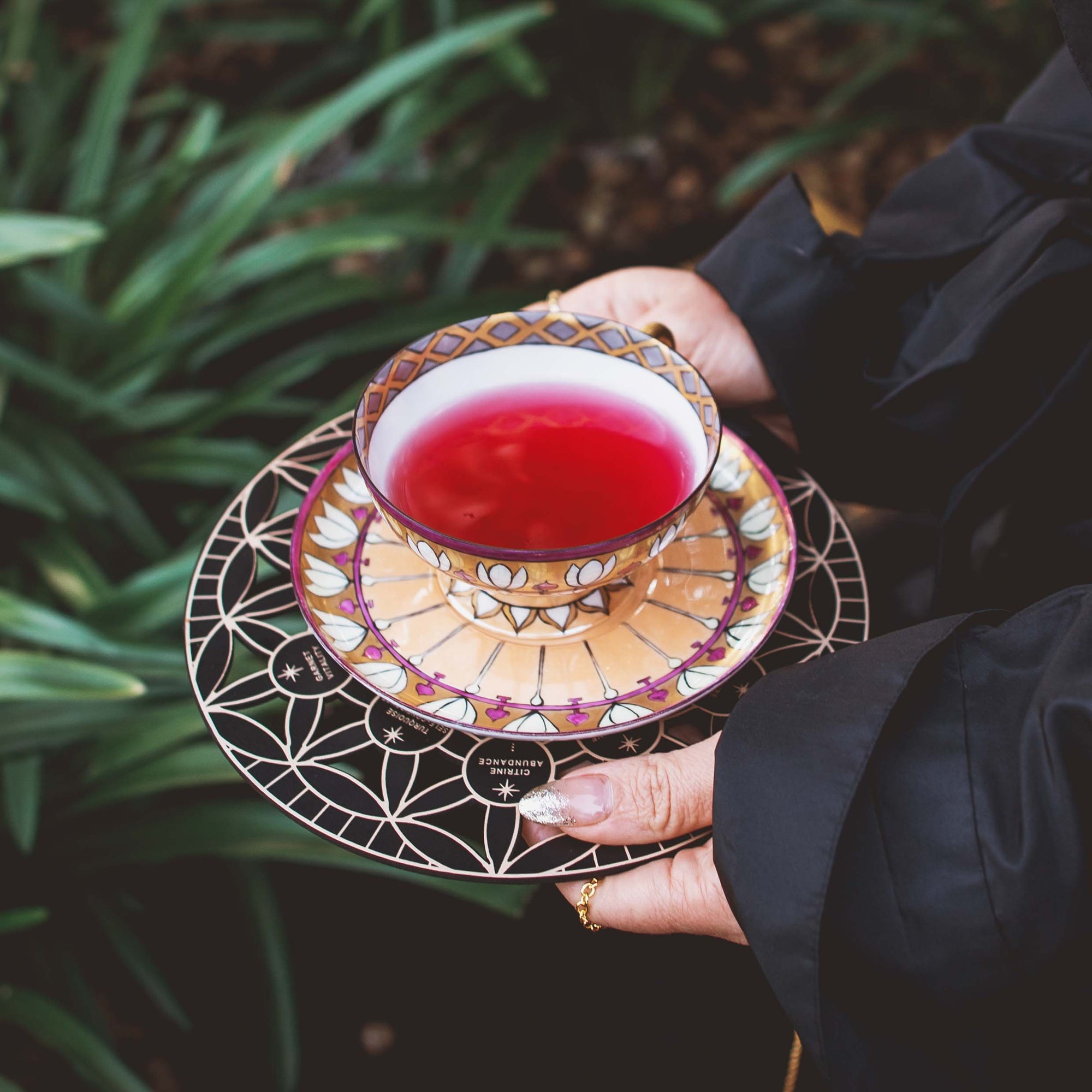 Someone in a dark sleeve holds a decorative teacup filled with red tea on a matching saucer, with green leafy plants and the Crystal Grid: Flower of Life Tea Ceremony Altar softly visible in the blurred background.