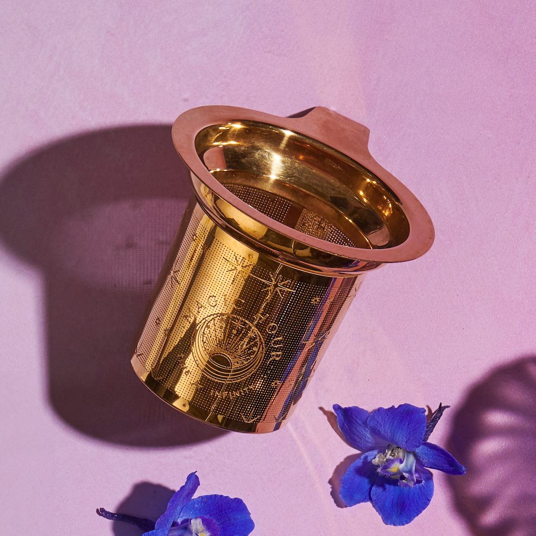 A gold-toned Steep The Stars - Artisan Tea Strainer by Magic Hour with intricate patterns is placed on a pink surface. Next to it, a small, vibrant blue flower petal adds a touch of color. The shadow of the Steep The Stars - Artisan Tea Strainer is cast on the surface, enhancing the image's depth—a perfect setting for brewing your favorite loose leaf tea.