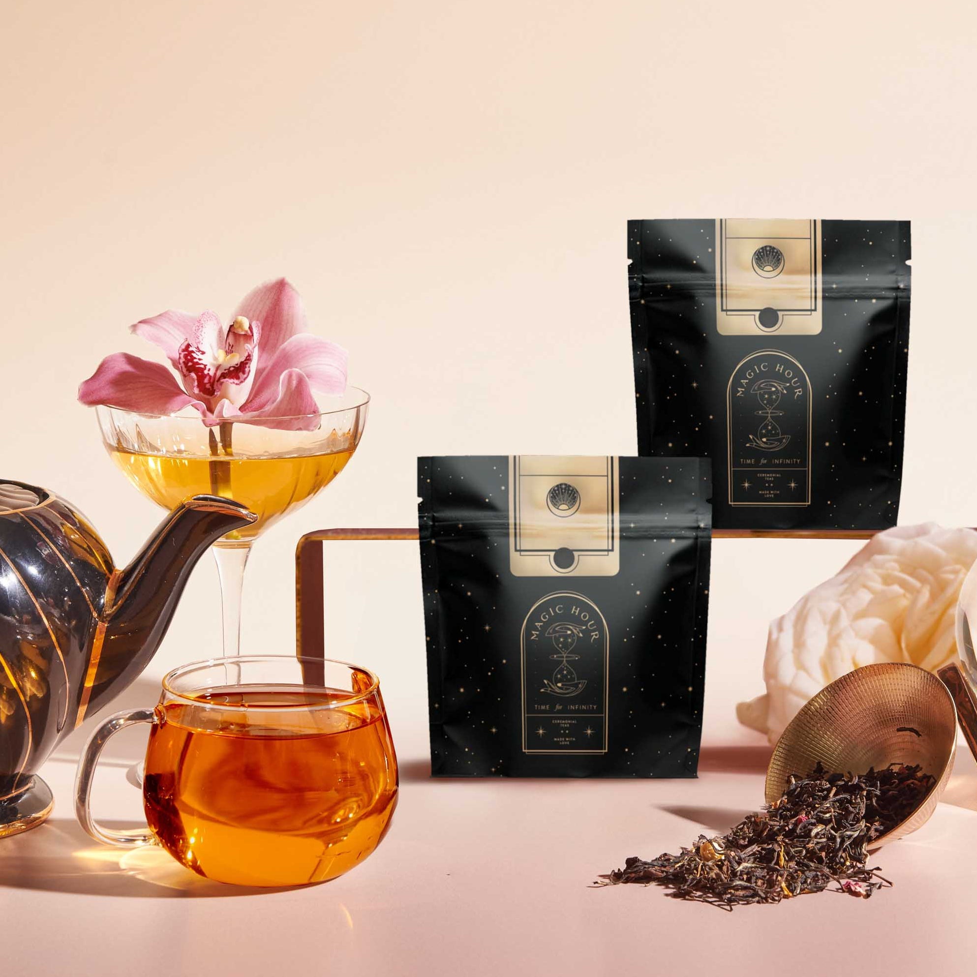 A black and gold teapot, glass cup of tea, cocktail glass with a pink orchid, two black tea packages, and loose tea leaves are artfully arranged to highlight the Tea Discovery Club sampler on a light background.