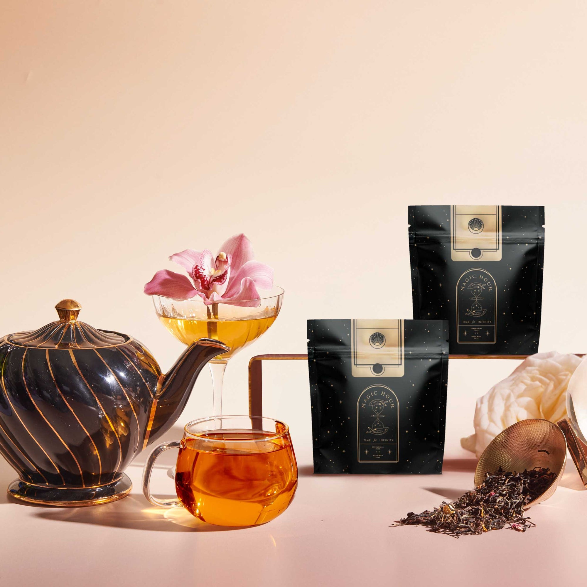 A black and gold teapot, glass cup of tea, cocktail glass with a pink orchid, two black tea packages, and loose tea leaves are artfully arranged to highlight the Tea Discovery Club sampler on a light background.