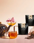 A black and gold teapot, glass cup of tea, cocktail glass with a pink orchid, two black tea packages, and loose tea leaves are artfully arranged to highlight the Tea Discovery Club sampler on a light background.