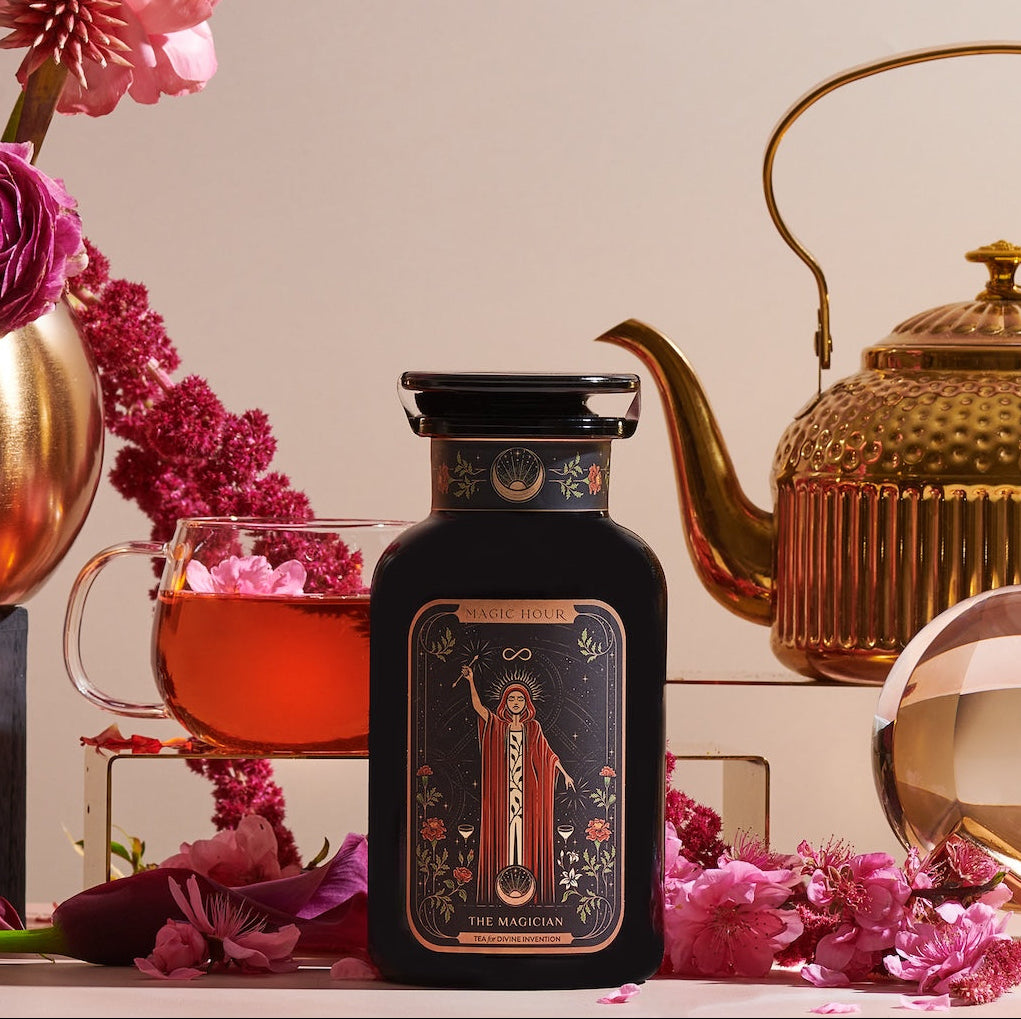 A black bottle labeled "The Magician," evoking transformative abilities, is surrounded by pink flowers, a glass cup of tea, a golden teapot, and a glass orb. The background features more pink flowers and a light-colored backdrop, creating a luxurious and elegant setting with wellness benefits.