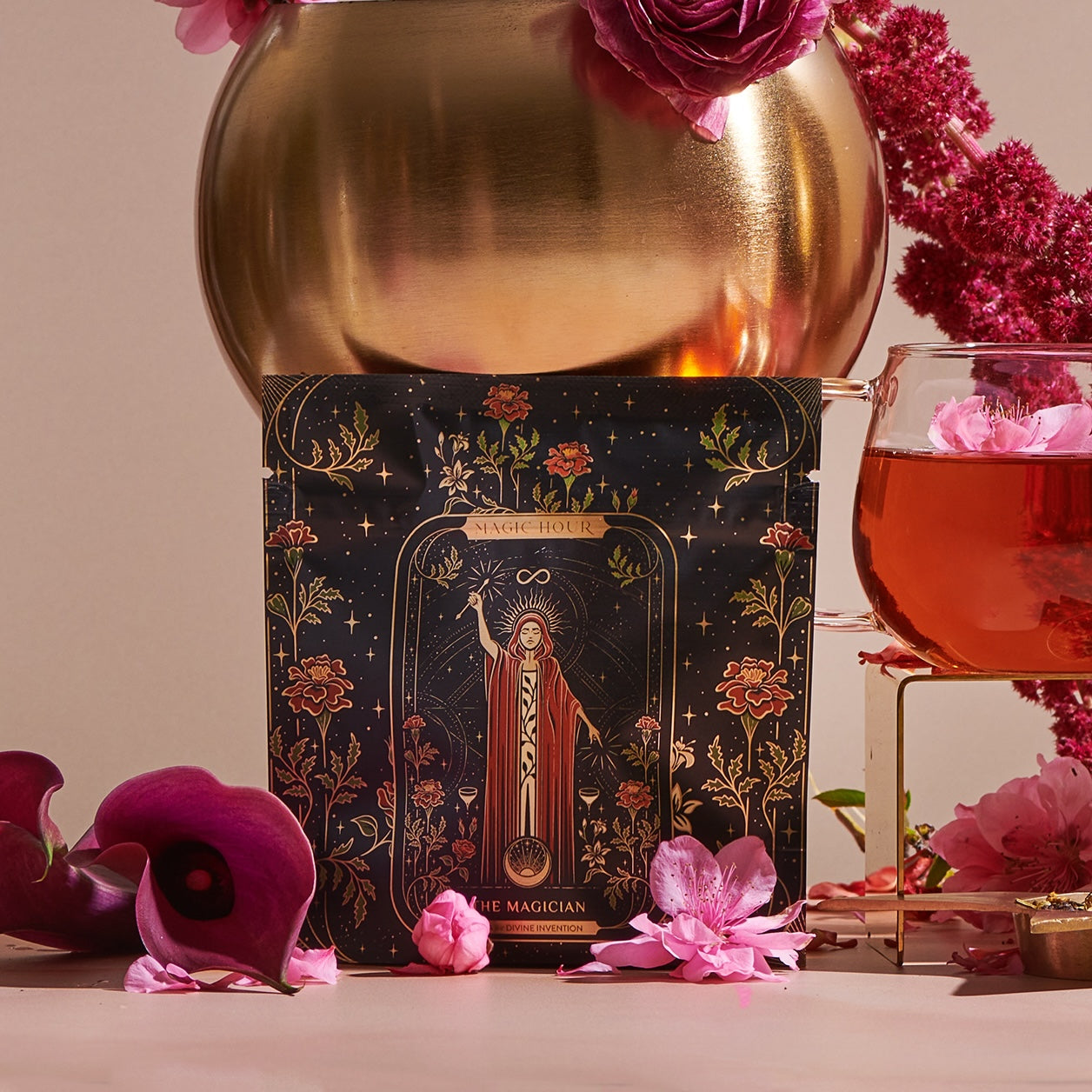 A colorful tarot card, "The Magician" from Magic Hour, showcasing transformative abilities, is displayed partially inside a golden vase adorned with pink roses. There is a glass of pink liquid with flower petals nearby, surrounded by various vibrant flowers on a soft pink background.