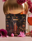 A 12 Tarot Tea Sampler Bundle card rests against a golden vase with flowers. A clear cup with pink tea and petal garnish hints at a Tarot Tasting Experience, while surrounding orchids and blossoms add to the enchanting vibe.