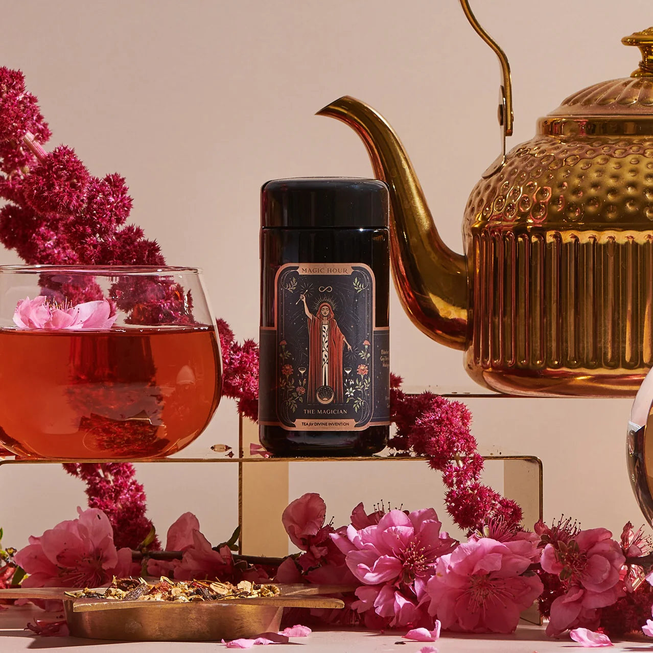 A decorative tea scene showcases a glass teacup with red tea, a jar labeled "The Magician," and a golden teapot amid pink flowers. Enhanced by violet glass jars and herbs, it exudes an elegant atmosphere reminiscent of the 12 Tarot Tea Traveler Jar Bundle.