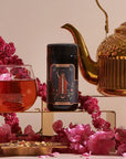 A decorative tea scene showcases a glass teacup with red tea, a jar labeled "The Magician," and a golden teapot amid pink flowers. Enhanced by violet glass jars and herbs, it exudes an elegant atmosphere reminiscent of the 12 Tarot Tea Traveler Jar Bundle.