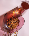 A heart-shaped, translucent pink bowl filled with loose leaf tea and a silver spoon rests on a surface. Next to the bowl is a clear glass pitcher, partially filled with red liquid, casting a shadow on the surface. The scene is lit with warm, diffuse light, invoking the transformative abilities of Magic Hour's The Magician wellness benefits.