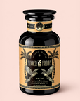 A black bottle with gold accents, palm trees, and cockatoos holds 20 plant-based sachets of “Mai Tai: Black & Puerh Tropical Mocktail Tea for Energizing Your Season of Sunshine.” This summer-inspired tea blend features notes reminiscent of Mai Tai cocktails and Puerh black tea. The set is displayed against a pale pink background.