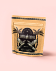 A tan resealable pouch labeled "Mai Tai: Black & Puerh Tropical Mocktail Tea for Energizing Your Season of Sunshine" features palm trees and birds in black and gold on a pink background. Contains 2.5 oz of energizing summer tea.