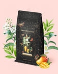 A Mango Jasmine Oolong Iced Tea bag, adorned with floral and butterfly motifs, is artfully displayed amid jasmine blossoms, lush green leaves, and fresh mango slices on a soft pink backdrop. The label reads "Mango Jasmine Oolong Iced Tea." This visually captivating tea offers a refreshing and uplifting mood-enhancing experience.