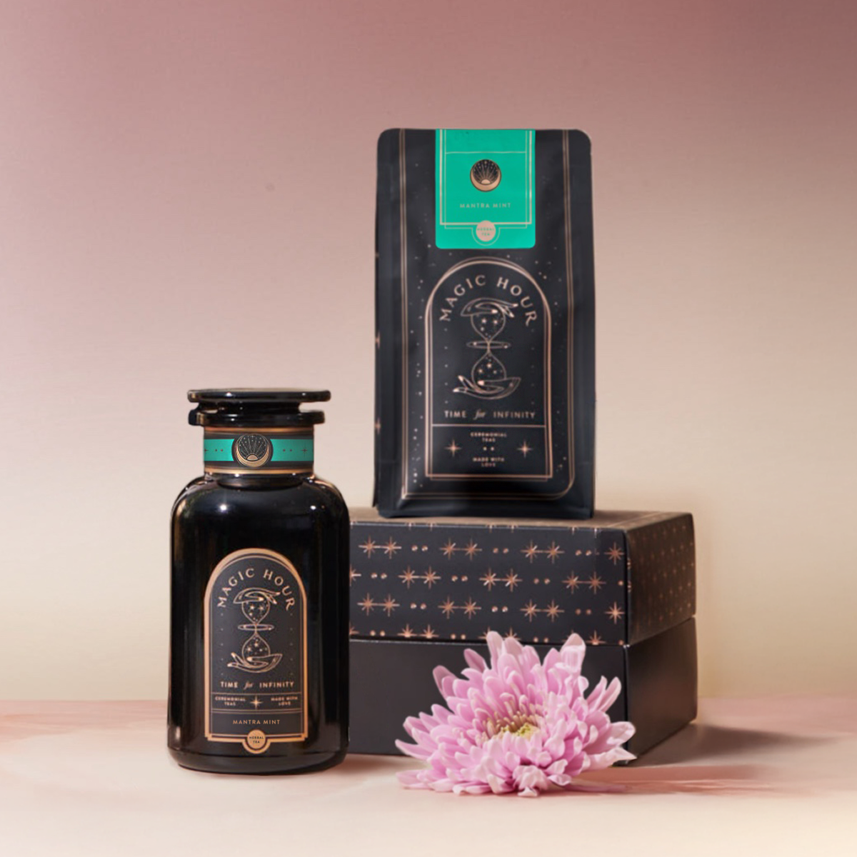The Mantra Mint™ Herbal Tea Starter Kit, featuring "Matcha Mint" jars and bags, is displayed with a pink chrysanthemum on a decorative box against a pink ombre backdrop. This caffeine-free blend is showcased beautifully.