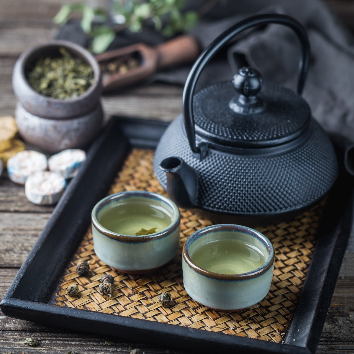 A black cast iron teapot sits on a woven mat placed on a wooden tray, accompanied by two small ceramic cups filled with premium Mao Feng Yunnan Green Tea from Magic Hour. In the background, a small wooden scoop and a container with loose tea leaves can also be seen.