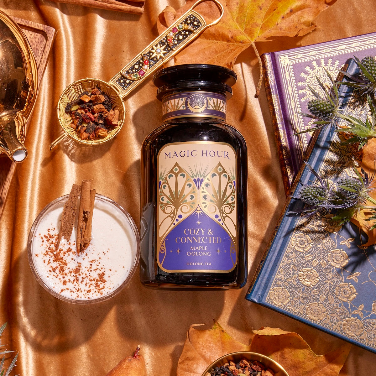 A bottle of Maple Oolong tea is tastefully arranged amidst autumn-themed items such as a gold pot, a cup of milk with cinnamon sticks, bowls of dried herbs, a decorative spoon, and an intricately designed open book. The golden fabric backdrop perfectly enhances the warm autumn flavors.