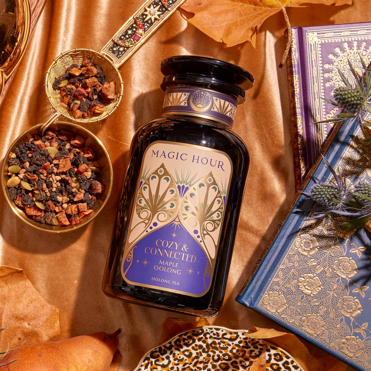 A decorative black bottle labeled "Magic Hour Cozy & Connected Maple Oolong" is placed on a satin cloth. Surrounding the bottle are a gold tea infuser, a bowl of tea leaves, autumn leaves, and ornate blue and gold books. The setting exudes a warm ambiance with rich autumn flavors.