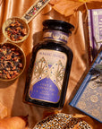 A decorative black bottle labeled "Magic Hour Cozy & Connected Maple Oolong" is placed on a satin cloth. Surrounding the bottle are a gold tea infuser, a bowl of tea leaves, autumn leaves, and ornate blue and gold books. The setting exudes a warm ambiance with rich autumn flavors.