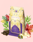 A decorative package of Maple Oolong: Cozy & Connected tea—a metabolism-boosting blend—is surrounded by loose tea leaves, red and green leaves, and apple slices on a light pink background, evoking the cozy charm of an autumn tea blend.