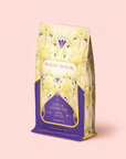 A decorative Maple Oolong Tea Refill Pouch stands upright on a light pink background, showcasing a yellow, purple, and teal art deco design. This energizing tea is an oolong apple blend crafted to delight your senses and support your morning ritual.