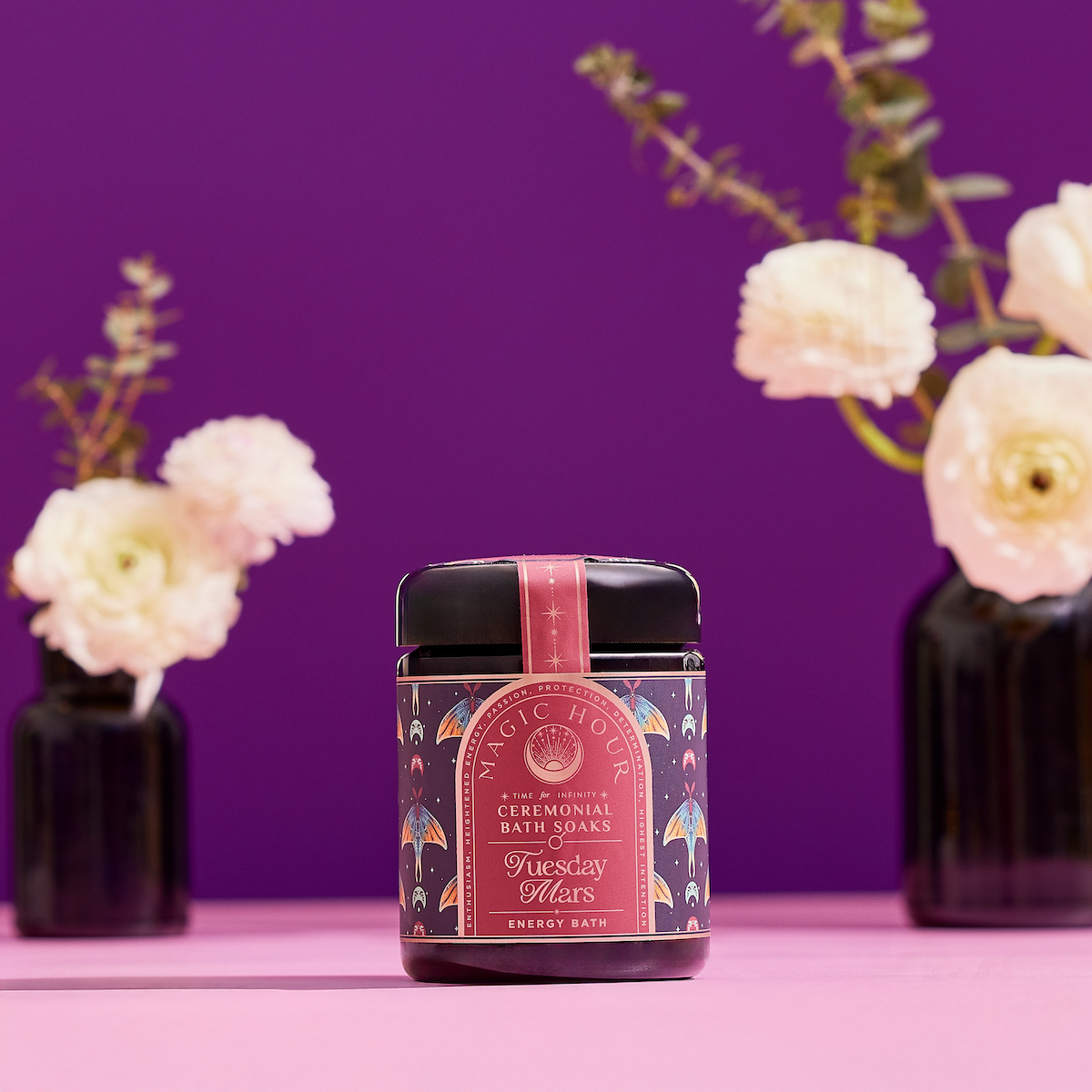 A decorative jar of &quot;Ceremonial Bath Salt Soaks&quot; in a violet glass jar rests on a pink surface, with blurred white flowers and vases against a purple backdrop—an elegant addition to your next self-care ritual.