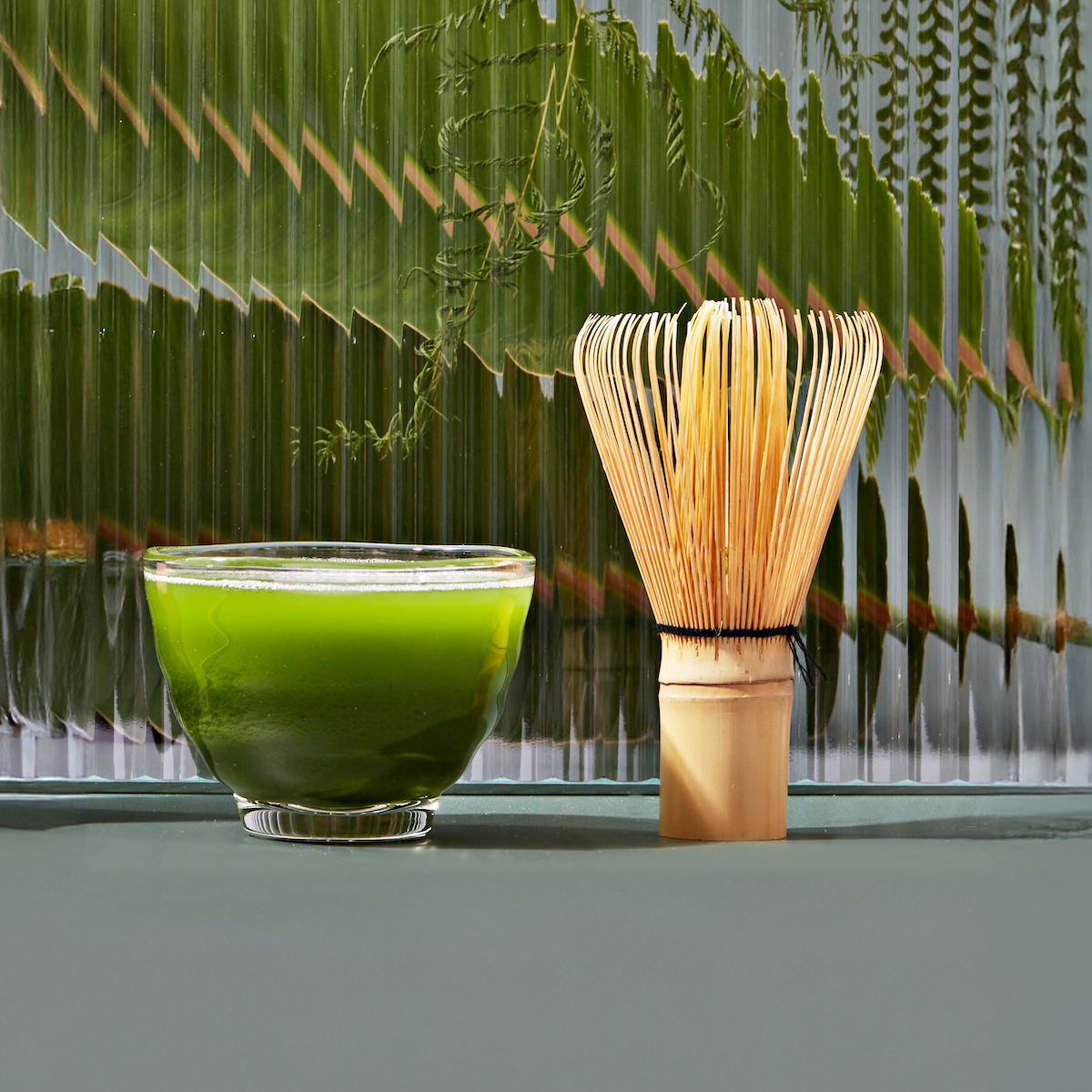 A bowl of Culinary Matcha 500 is beside a bamboo whisk on a green surface, with large tropical leaves in the background, creating a fresh and natural atmosphere.