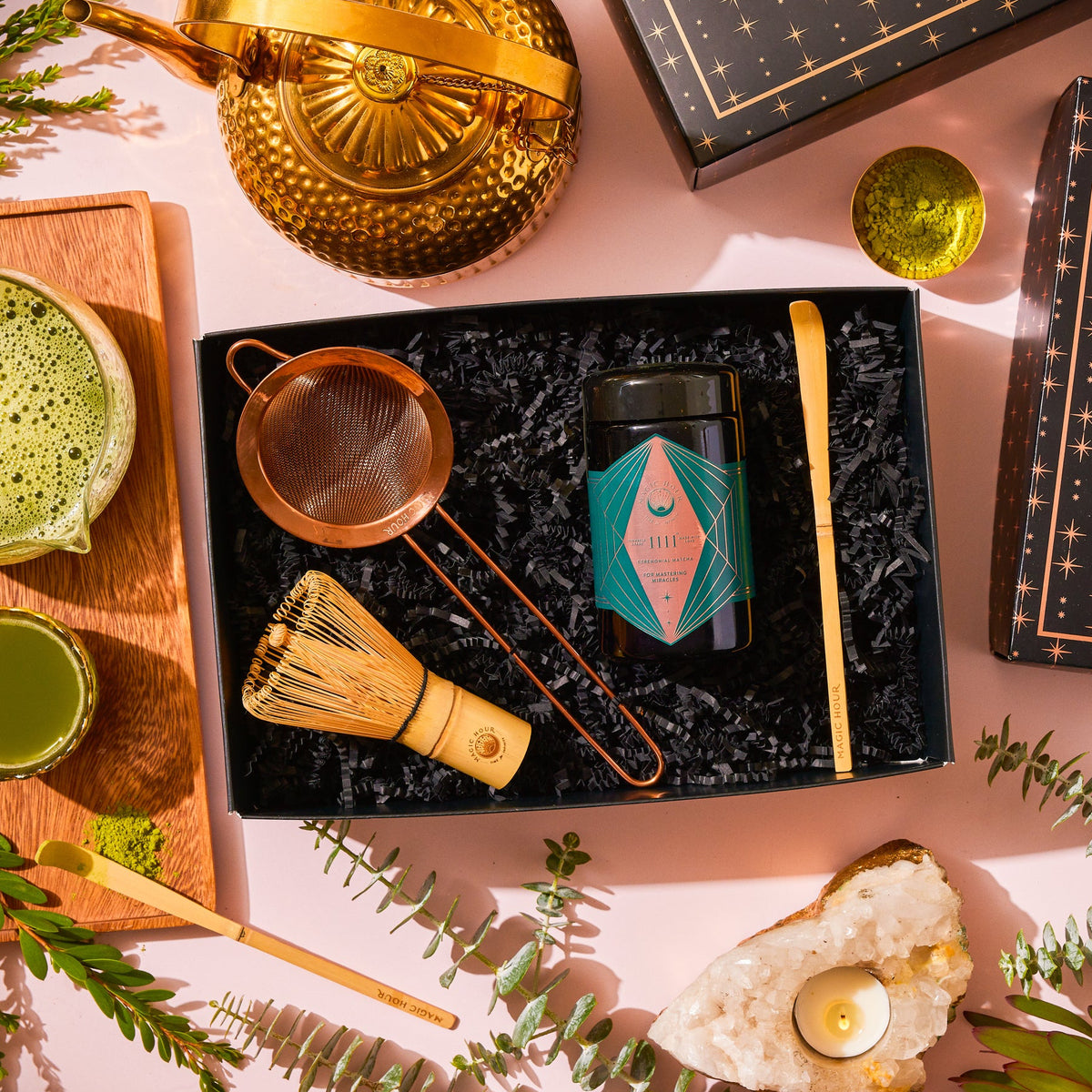 A Matcha 1111 : Gift Set containing a bamboo matcha whisk, powder canister, strainer, and scoop, arranged with a gold teapot, a bowl of prepared Japanese matcha tea, and a crystal candle holder.