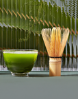 A bowl of Culinary Matcha 500 is beside a bamboo whisk on a green surface, with large tropical leaves in the background, creating a fresh and natural atmosphere.