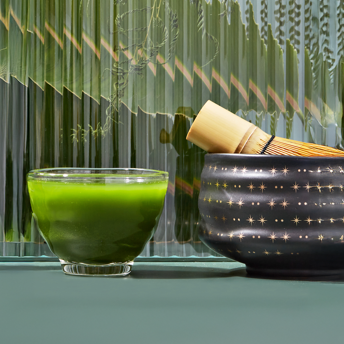 A glass bowl filled with vibrant green Culinary Matcha 500 sits beside a black ceramic bowl adorned with golden star patterns, holding a bamboo whisk. The background showcases textured green glass panels.