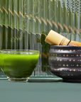 A glass bowl filled with vibrant green Culinary Matcha 500 sits beside a black ceramic bowl adorned with golden star patterns, holding a bamboo whisk. The background showcases textured green glass panels.