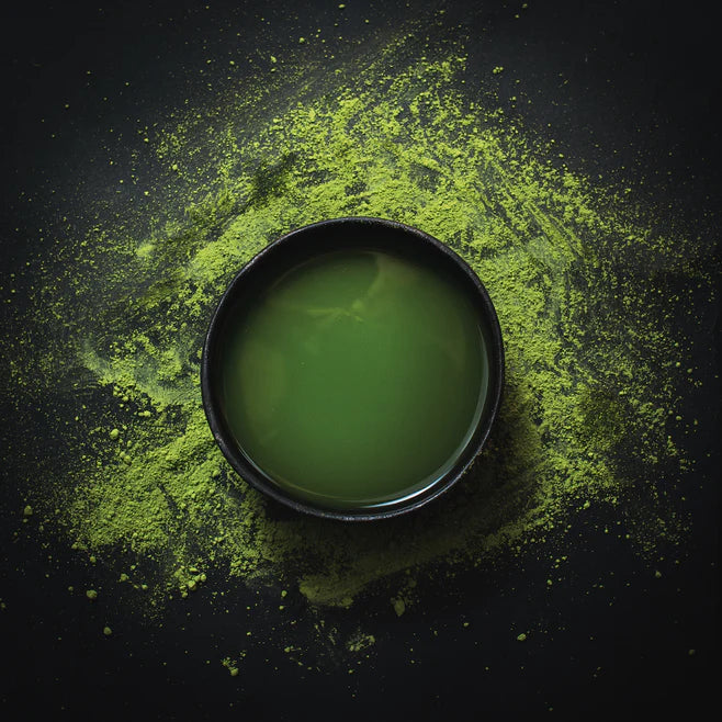 A dark bowl filled with vibrant green liquid sits atop a scattered mound of matcha powder, against a dark background. The arrangement highlights the vivid color and texture contrast between the fine powder and the smooth liquid.