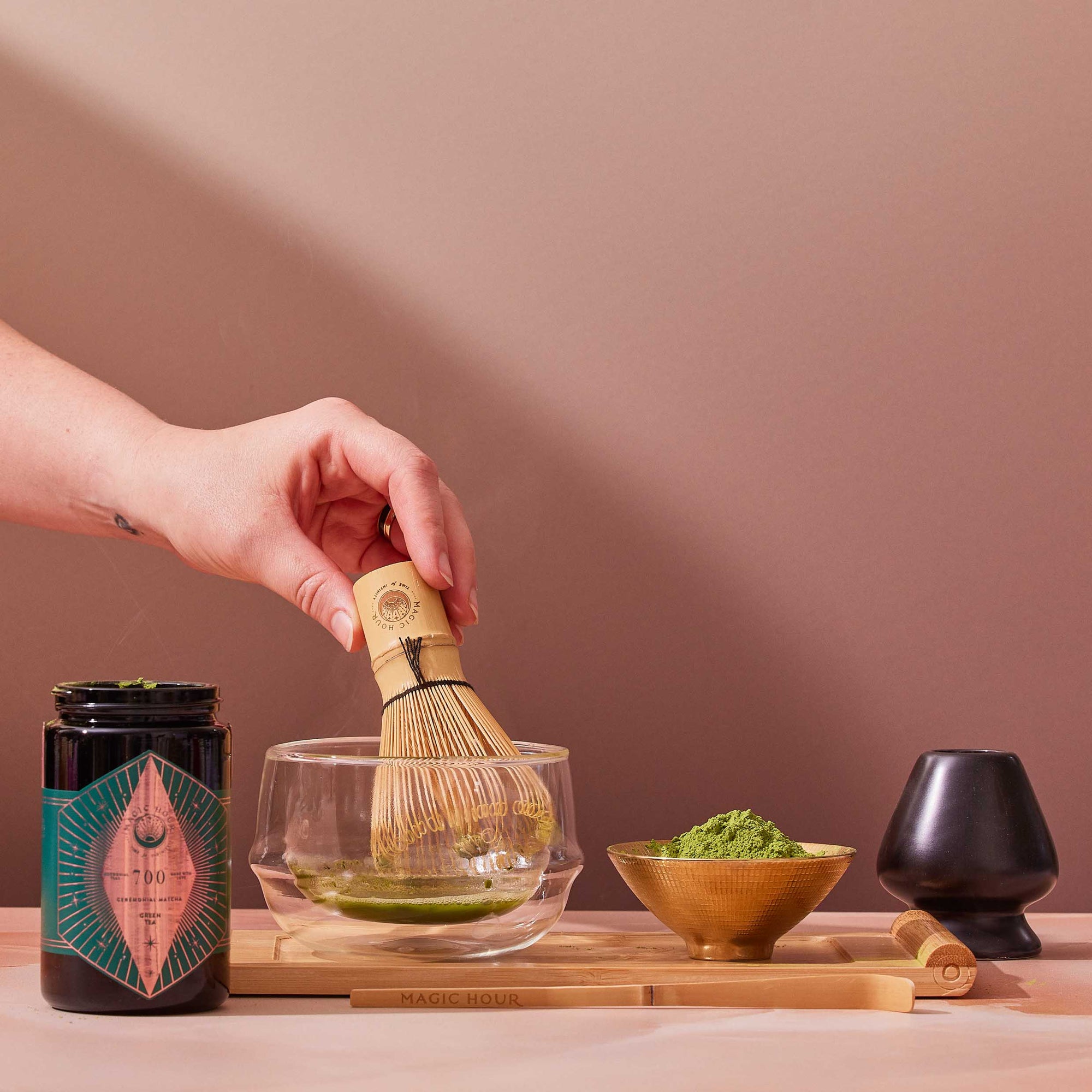 A bamboo whisk stirs Organic Ceremonial Matcha 700 in a glass bowl. On a wooden tray are a jar of matcha powder, a bowl with matcha, and a small black cup.