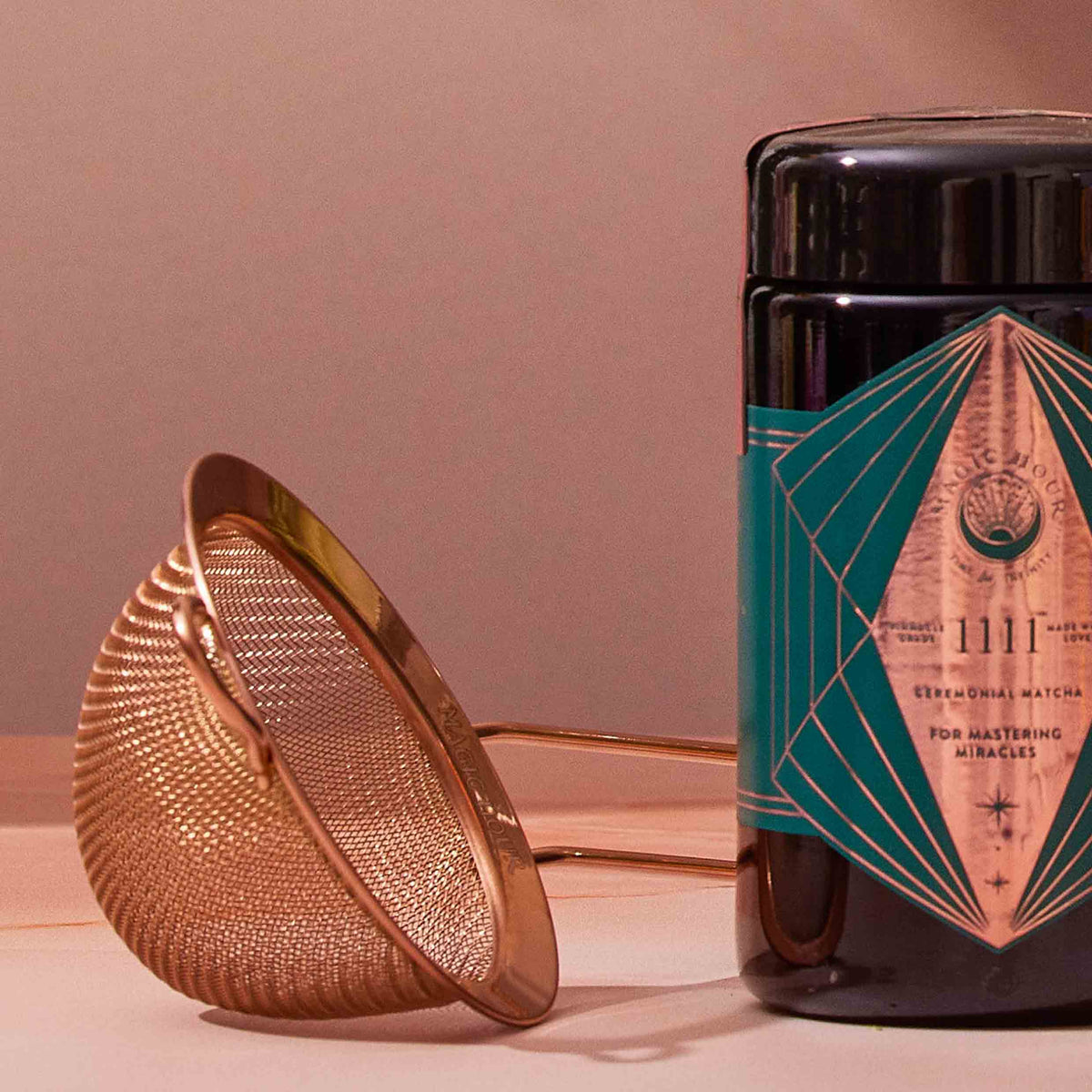 A Rose Gold Matcha Sift rests beside a black jar adorned with a turquoise and copper geometric label on a wooden surface, ready to elevate your matcha experience.