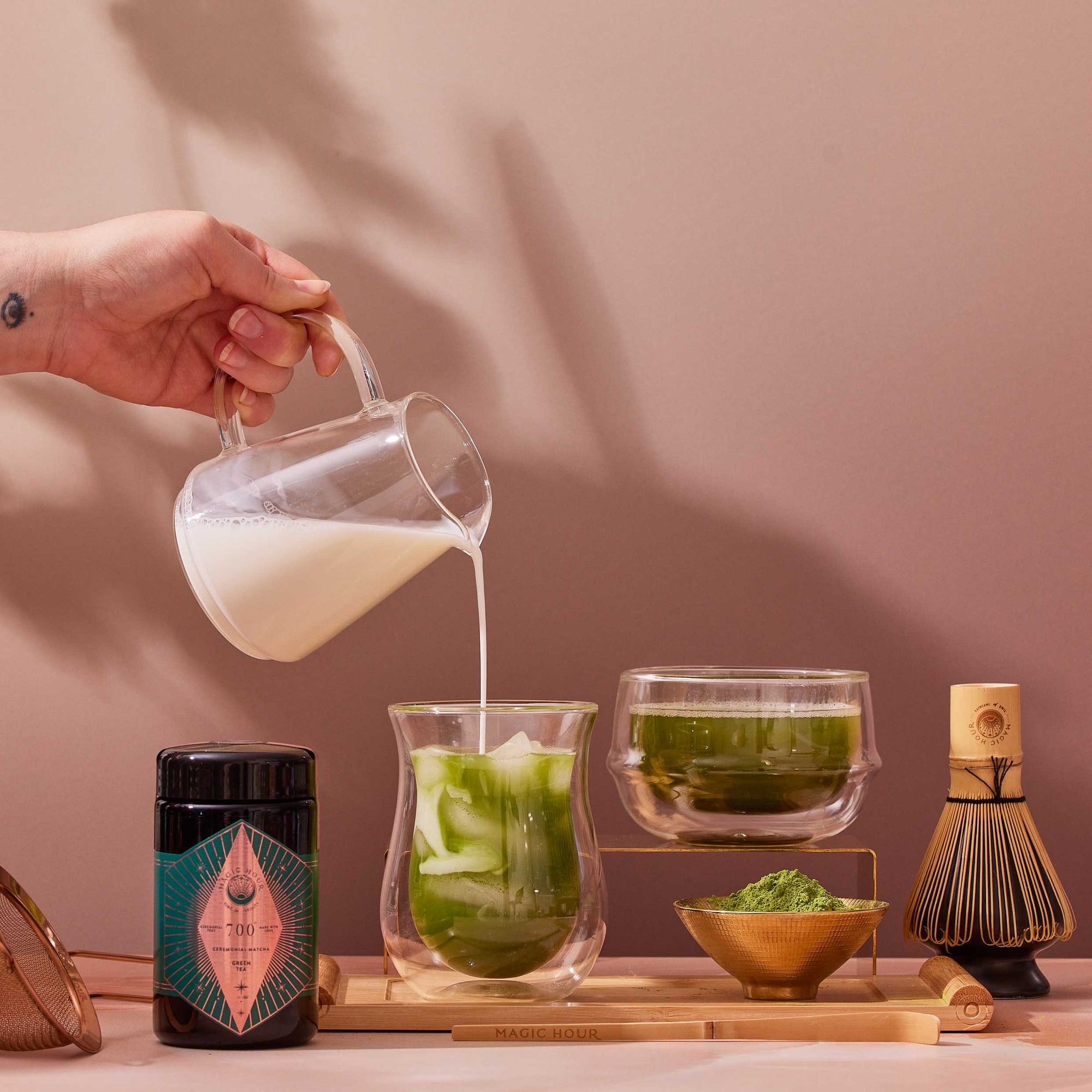 A hand pours milk into a glass of iced Kagoshima matcha. Nearby are a jar of Organic Ceremonial Matcha 700 powder, a bowl of tea, a bamboo whisk, a scoop with green powder, and a fine mesh strainer on a tray.